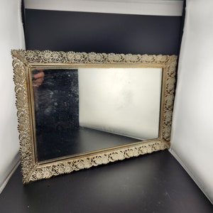 May include: A gold-colored metal tray with a mirrored surface and a decorative floral design around the edge.
