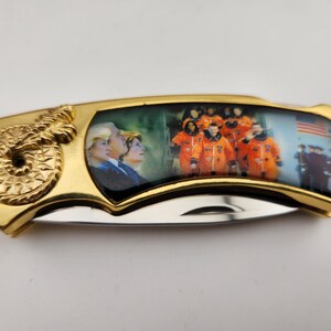 May include: A gold pocket knife with a picture of astronauts in orange space suits and a picture of a man and woman on the other side.