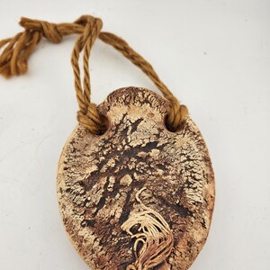 May include: A small, brown, ceramic, heart-shaped pouch with a knotted rope handle. The pouch has a textured surface and a design of a stylized tree with a single branch.