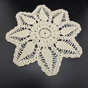 May include: A round, off-white crochet doily featuring a central circular pattern surrounded by eight radiating petal-like shapes. The design incorporates intricate openwork stitching and delicate thread loops that create a star-like silhouette.