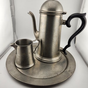 May include: A pewter tea set with a teapot, creamer, and tray. The teapot has a black handle and a lid with a finial. The creamer has a small handle. The tray is round and has a raised edge.