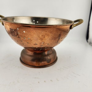 May include: A copper colander with brass handles and a stand. The colander has a round shape and is perforated with small holes.