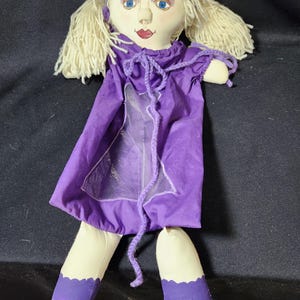 May include: A handmade rag doll with blonde yarn hair, blue eyes, and a purple dress. The doll has a white face and red lips. The doll is wearing black shoes with purple trim.
