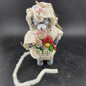 May include: A gray mouse figurine wearing a white lace bonnet with a pink bow and a matching dress. The mouse holds a small wreath with a red bow and a gold bell. A long, white, fuzzy tail extends from the mouse.