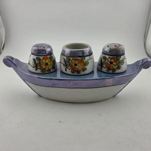 May include: A ceramic salt and pepper set with a matching toothpick holder. The set is shaped like a boat, with a lavender rim and handles. The salt and pepper shakers and toothpick holder are white with floral designs.