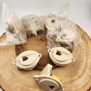 May include: Several off-white electrical components, some in plastic packaging, are arranged on a wooden surface. The components have a circular design with a square opening and small wires. The wood has a natural, light brown color.