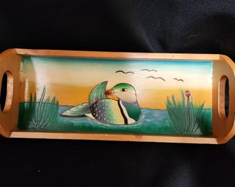 May include: A wooden serving tray with a painted scene of a duck swimming in a pond with green reeds and a pink flower. The duck is green, blue, and yellow.