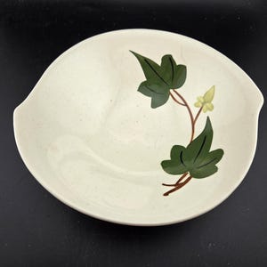 May include: Cream-colored ceramic bowl with a speckled finish and two handles. The bowl is decorated with a green ivy design and a yellow flower. A vintage piece for serving.