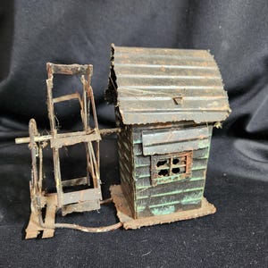 May include: A rustic, miniature water mill model. The structure features a small wooden house with a corrugated metal roof and a water wheel. The model is painted in shades of green and brown, with a weathered appearance.