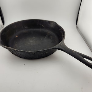 May include: A black cast iron skillet with a rough, textured surface. The skillet has a long handle with a loop at the end.