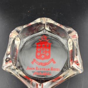 May include: A clear glass ashtray with a red logo that says "John Bartram Hotel Philadelphia" and a crest with a red "G" and red crosses.