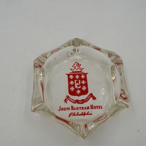 May include: Clear glass ashtray with a white center featuring a red crest and the text "JOHN BARTRAM HOTEL Philadelphia". The ashtray has a hexagonal shape with six indentations for resting cigarettes.