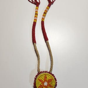 May include: A beaded necklace with a round pendant featuring a yellow, orange, and white flower design. The necklace is made with red, yellow, and white beads and has a brown cord.