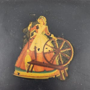May include: A vintage black box with a colorful image of a woman spinning a wheel. The woman is wearing a yellow dress and a red bodice. The wheel is brown with gold accents.