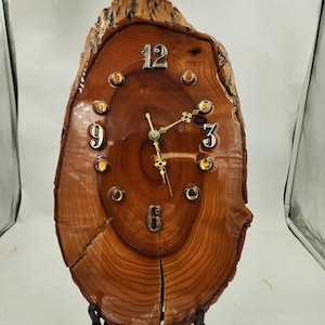 May include: A wooden clock with a natural wood finish and a round face. The clock has a metal stand and features Roman numeral hour markers and gold-colored hands.