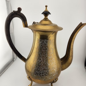 May include: A brass teapot with a wooden handle and an ornate design. The teapot has three legs and a lid with a black knob.