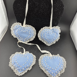May include: Four light blue heart-shaped ornaments with white lace trim and white dotted accents. Each heart has a white ribbon for hanging. The ornaments are displayed against a black background.