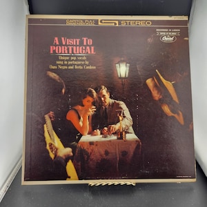 May include: Vintage vinyl record album cover titled "A Visit To Portugal." The cover features a romantic scene with a couple at a table, a musician playing a guitar, and the text "Unique pop vocals sung in portuguese by Ouro Negro and Berta Cardoso."