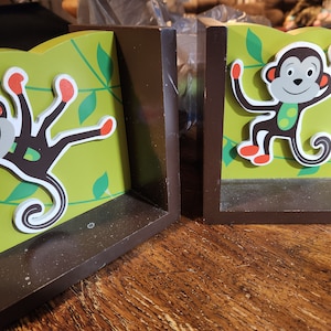 May include: A pair of brown wooden bookends with a green background and a cartoon monkey design. One monkey is hanging from a vine and the other is standing on a vine.