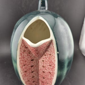 May include: A ceramic pitcher shaped like a watermelon. The exterior is a dark green, with a red and white interior. The pitcher has a handle and a spout.