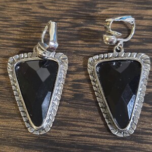 May include: A pair of silver-toned clip-on earrings. Each earring features a black, triangular-shaped stone set within a decorative silver frame. The frame has a textured, rope-like design around the edges. The earrings are displayed on a wooden surface.