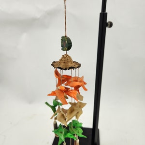 May include: A colorful wind chime with a woven hat and multiple layers of orange, brown, and green paper fish. The chime is hanging from a black metal stand.