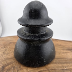 May include: A black, antique-style insulator, likely made of ceramic or glass. The insulator has a rounded top, a mid-section, and a flared base. It sits on a wooden surface, showcasing its vintage aesthetic. The object is a dark, solid color.