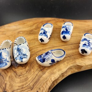 May include: Five miniature blue and white ceramic Dutch clogs with windmill and boat designs. The clogs are arranged on a wooden surface.
