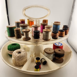 May include: A white plastic two-tiered sewing thread organizer with a handle. The top tier has 24 spools of thread in various colors, including red, blue, green, pink, and brown. The bottom tier has four compartments with spools of thread and yarn.