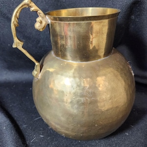 May include: A brass pitcher with a hammered finish and a decorative handle. The pitcher has a round body and a wide mouth.