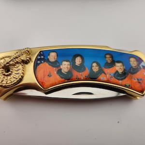 May include: A gold pocket knife with a blue and white image of astronauts in orange space suits. The knife has a decorative gold handle with a coiled design.