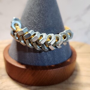 May include: A silver and gold bracelet made of nuts and bolts. The bracelet is displayed on a gray cone-shaped stand with a wooden base.