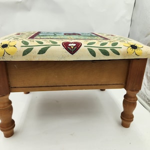 May include: A wooden footstool with a floral patterned fabric top. The fabric features yellow flowers, green leaves, and a red heart with a white flower inside. The stool has turned wooden legs.