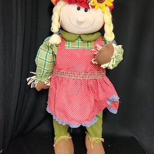 May include: A plush scarecrow doll with a red and white checkered dress, a green plaid shirt, and a brown hat with a yellow sunflower. The doll is holding a brown object in its right hand.