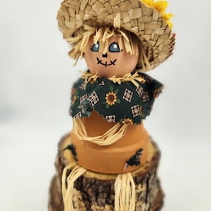 May include: A handmade scarecrow figurine made from a terracotta pot, straw, and fabric. The scarecrow is wearing a straw hat, a green and brown plaid fabric, and has a painted face with blue eyes. The scarecrow is sitting on a wooden log.