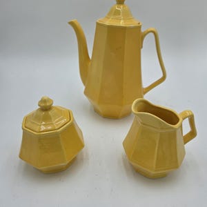 May include: A yellow ceramic tea set, including a teapot with a lid, a sugar bowl with a lid, and a creamer. The set has a geometric design and is set against a white background. The teapot is approximately 9 inches tall.