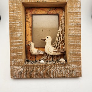 May include: A wooden picture frame with a nautical theme. The frame features a small picture window with a brown background, a net, and two white seagulls with brown accents. The frame is decorated with small white seashells and brown pebbles.