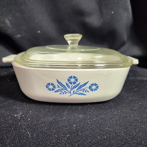 May include: White casserole dish with a glass lid and a blue floral design. The design features three blue flowers with green stems and leaves.