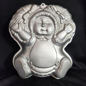 May include: A silver metal cake pan shaped like a Cabbage Patch Kid doll. The doll has pigtails, a dress, and shoes.