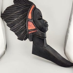 May include: A wooden wall sculpture of a person's head with a carved headdress. The sculpture is painted black with orange accents.