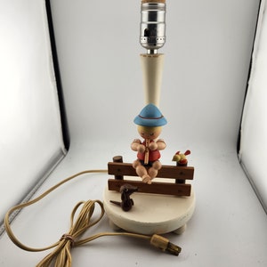 May include: A white table lamp with a wooden figurine of a boy playing a flute on a bench with a dog and bird. The lamp has a white cord.