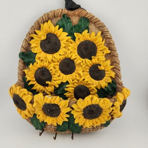 May include: A brown ceramic key holder shaped like a basket overflowing with yellow sunflowers and green leaves. A small black bird sits on the top of the basket.