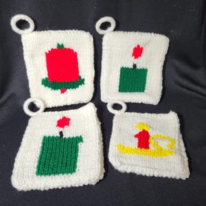May include: Four white knitted squares with different Christmas designs. Each square has a red, green, and yellow candle with a flame on top. The squares have a loop at the top for hanging.