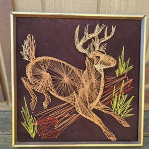 May include: A framed string art piece depicting a brown deer with antlers running through a field of green grass. The deer is made of string in shades of brown, orange, and yellow. The background is a dark brown felt.
