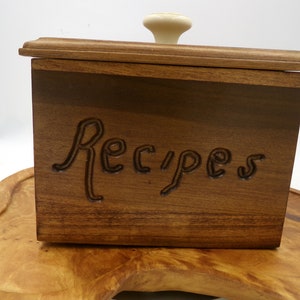 May include: A wooden recipe box with a white knob on the lid. The box is made of dark wood and has the word "Recipes" carved into the front.