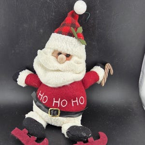 May include: A plush Santa Claus figure with a red and black plaid hat, white beard, and red suit. The figure has the words "HO HO HO" on the belly and holds a candy cane. Black boots and a black belt with a gold buckle complete the festive design.