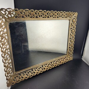 May include: A gold-colored metal tray with a decorative, ornate frame and a mirrored surface.