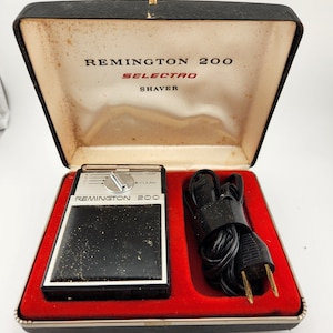 Może przedstawiać: Czarna elektryczna maszynka do golenia w czerwonym, aksamitnym etui z czarnym kablem. Maszynka do golenia jest oznaczona jako "Remington 200 Selecto Shaver" i ma pokrętło z ustawieniami "Trim" i "Clean".