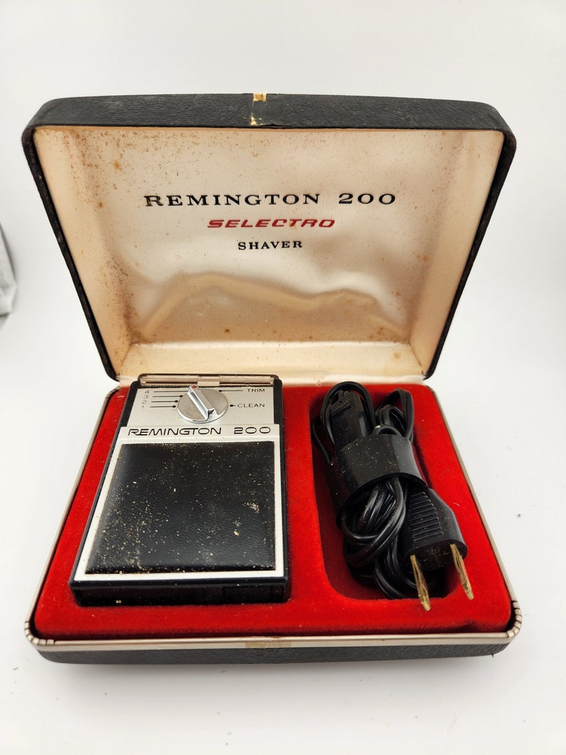 Może przedstawiać: Czarna elektryczna maszynka do golenia w czerwonym, aksamitnym etui z czarnym kablem. Maszynka do golenia jest oznaczona jako "Remington 200 Selecto Shaver" i ma pokrętło z ustawieniami "Trim" i "Clean".