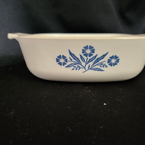 May include: A white rectangular casserole dish with blue floral design. The dish has two small handles and a slightly curved shape. The blue floral pattern includes three flowers and leaves. This vintage dish is suitable for baking and serving.